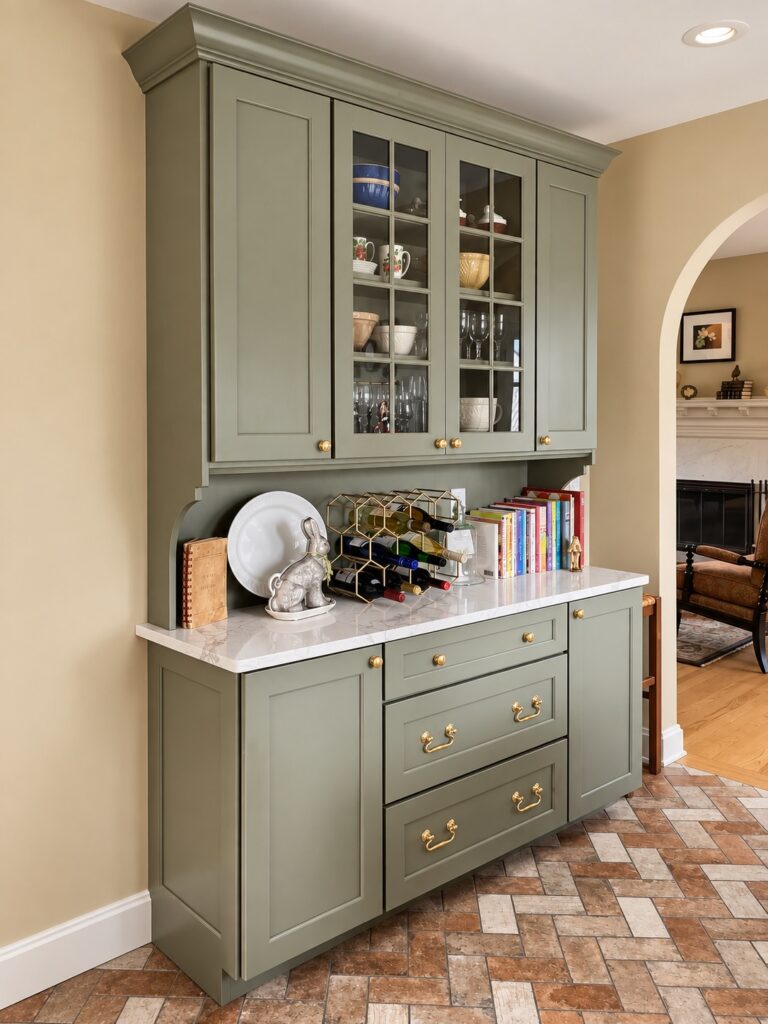 Sage green pantry hutch with glass-front upper cabinets, brass hardware and a white marble countertop beside a brick paver floor