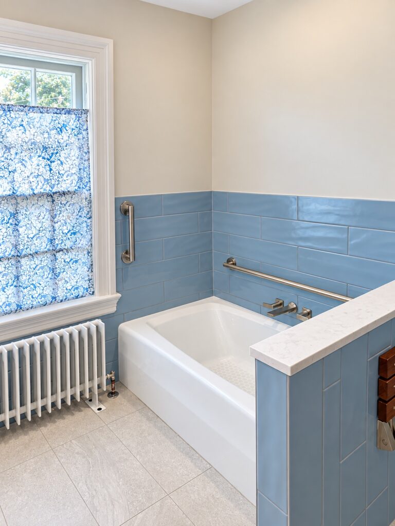 Blue tiled bathroom with a white soaking tub, brushed brass grab bars and a cast iron radiator beneath a stained glass window
