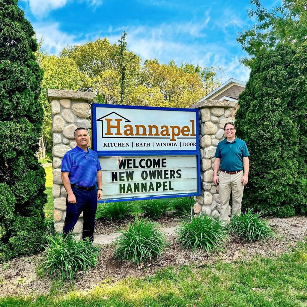 Two men standing beside a stone Hannapel showroom sign reading Welcome New Owners