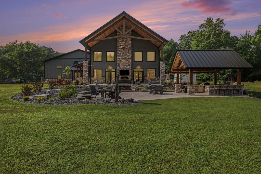 Custom KCH Homes build at dusk showing the rear exterior with stone and dark wood siding, a covered patio pavilion, outdoor seating, and a landscaped backyard