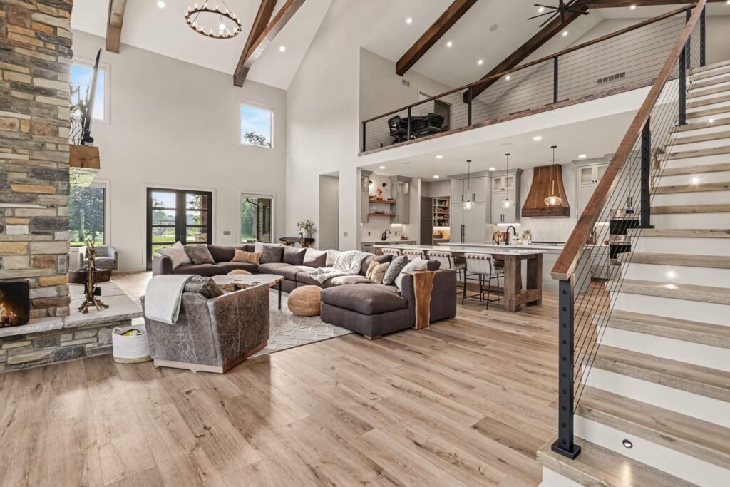 Open-concept great room and kitchen in a custom KCH Homes build with a floor-to-ceiling stone fireplace, exposed wood beams, cable-rail staircase, and hardwood flooring