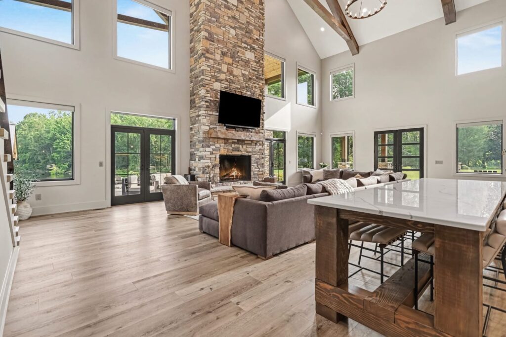 Vaulted great room in a custom KCH Homes build with a floor-to-ceiling stone fireplace, tall windows, exposed wood beam, hardwood floors, and a waterfall-edge kitchen island