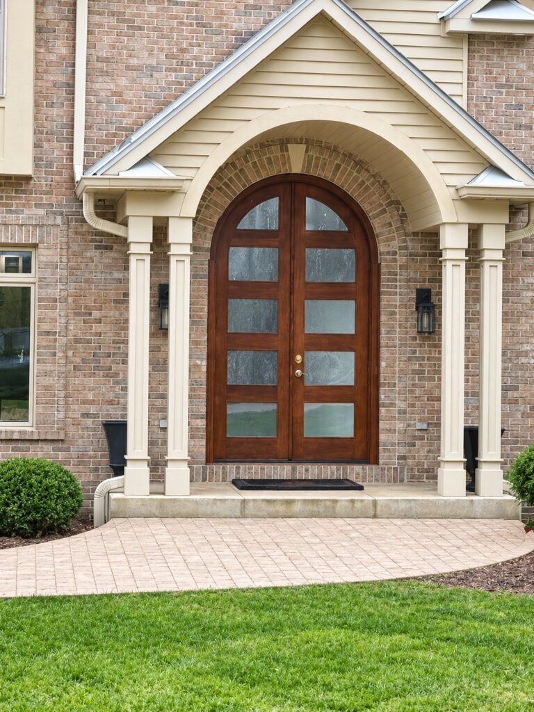 Finished custom arched Simpson double wood entry door with glass panels, surrounded by brick and a cream gabled porch.
