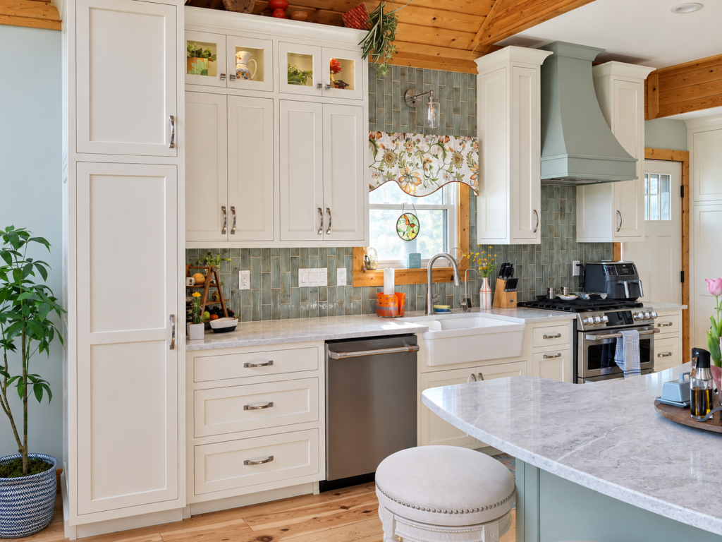 Cream pantry cabinets and glass-front uppers beside a farmhouse sink and sage range hood.