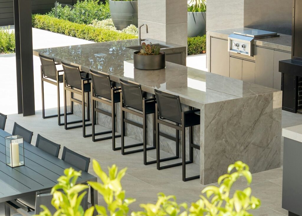 Covered outdoor kitchen with a veined Dekton island, bar seating and grill, by Cosentino