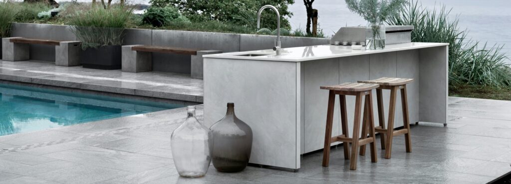 White Dekton outdoor kitchen island with stools beside a pool and lake, by Cosentino