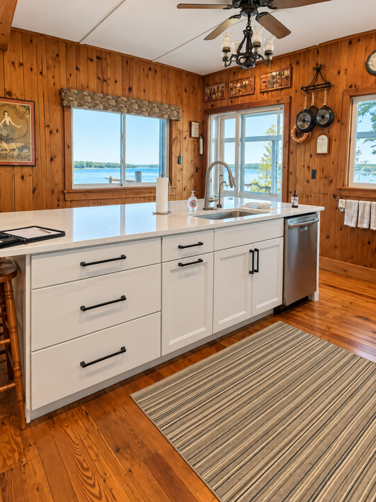 White sink cabinetry and stainless dishwasher beneath windows in a wood-lined kitchen.