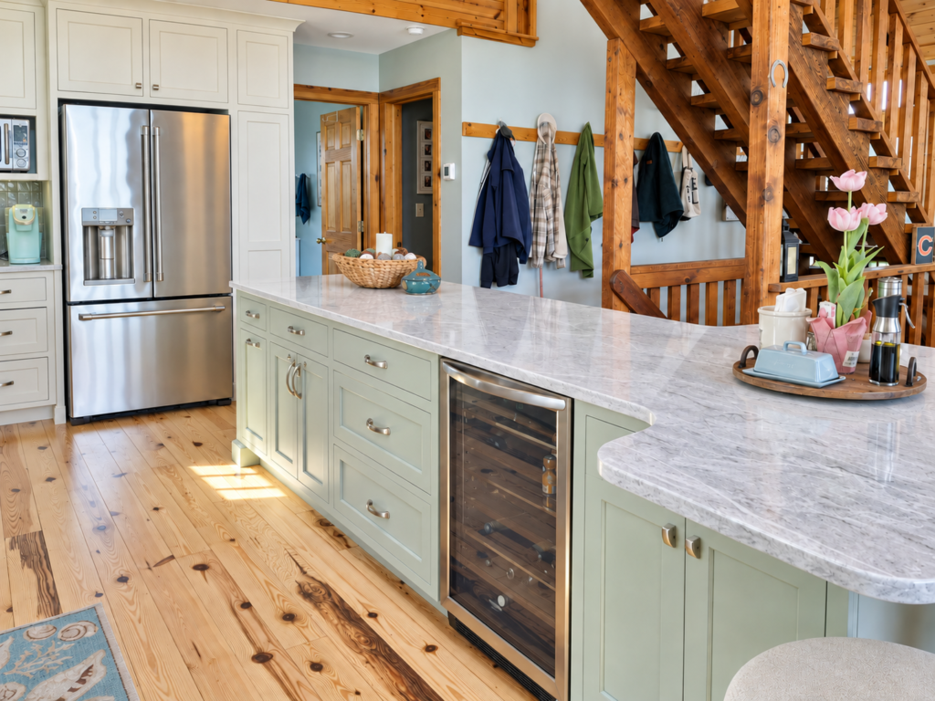 Sage kitchen island with a beverage refrigerator, pale stone countertop, cream cabinetry and wooden staircase beside a plain blue-gray wall.