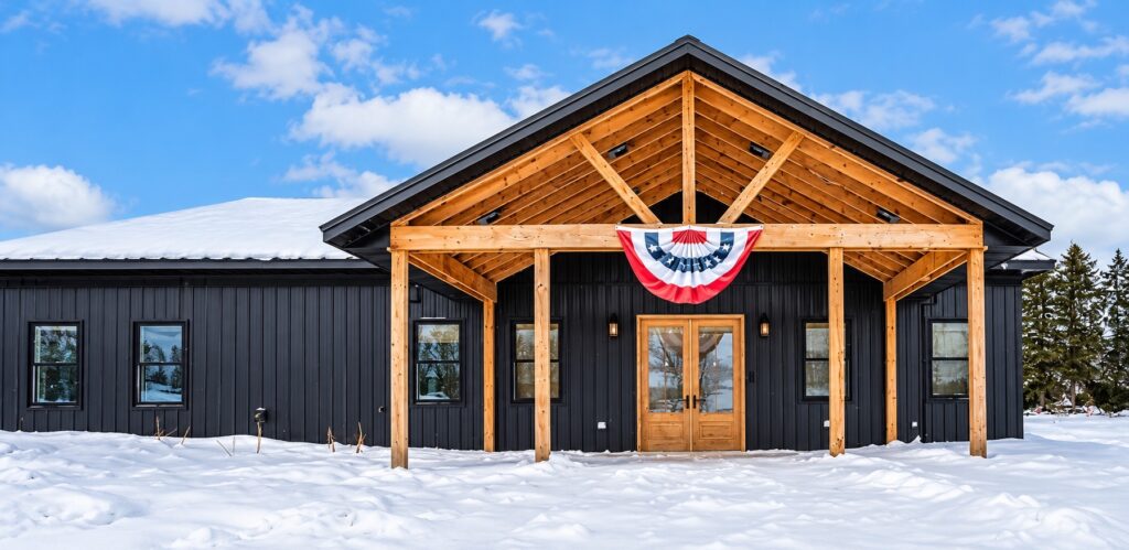 Daylight view of a black-sided home with a natural timber gabled porch and wood double entry doors in winter.