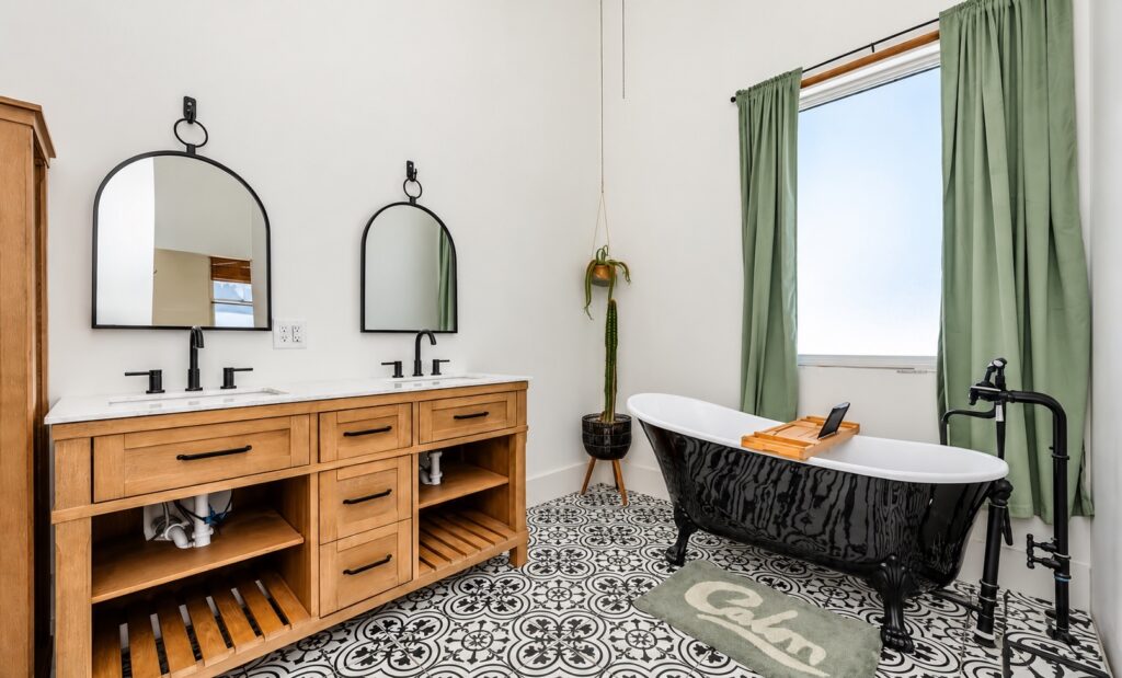 Bathroom with a natural wood double vanity, black arched mirrors, a black clawfoot tub, sage curtains and patterned tile.